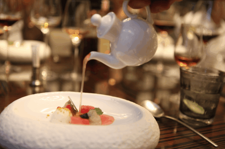 A server pours aromatic broth from a white teapot onto a meticulously plated dish featuring radish slices, mousse dollops, and edible flowers, set on a textured white plate in a refined dining setting. The elegant tablescape—complete with wine glasses and lime-garnished water—evokes the theatrical precision of Michelin dining, contrasting with the broader ambiance of fine dining