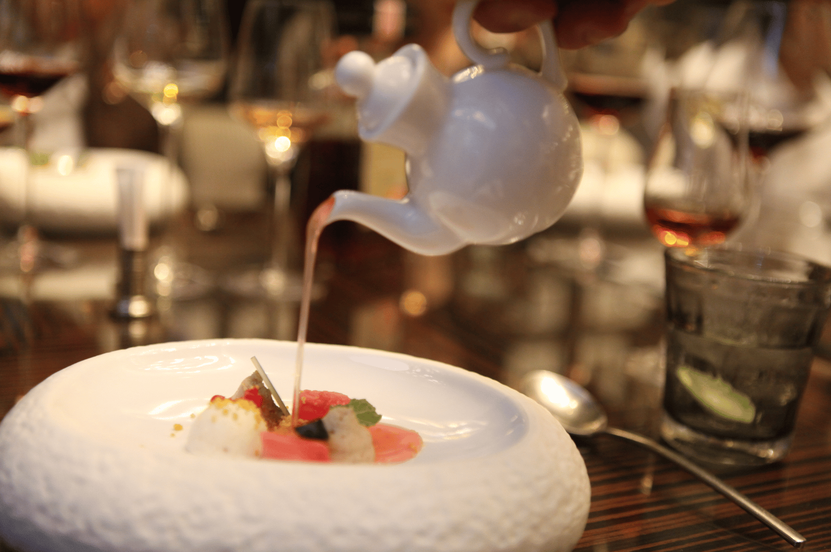 A server pours aromatic broth from a white teapot onto a meticulously plated dish featuring radish slices, mousse dollops, and edible flowers, set on a textured white plate in a refined dining setting. The elegant tablescape—complete with wine glasses and lime-garnished water—evokes the theatrical precision of Michelin dining, contrasting with the broader ambiance of fine dining