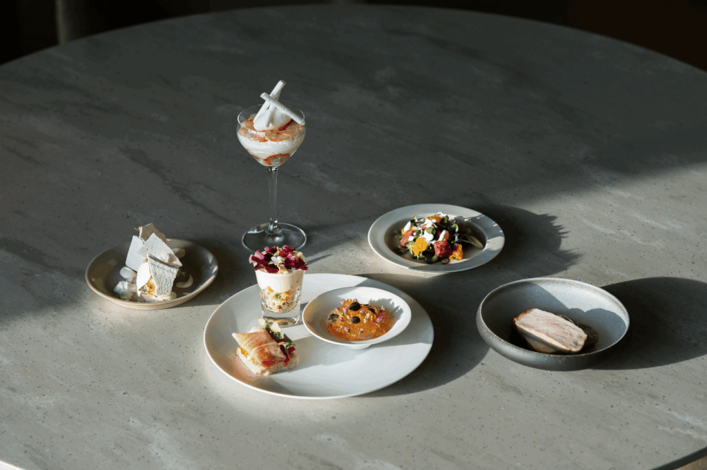 Six gourmet dishes arranged on a light-toned round table showcase the artistry of a tasting menu experience. From layered desserts with meringue to vibrant salads, seared meats, and edible flower garnishes, each plate offers a distinct visual and textural narrative—reflecting the creative freedom of fine dining, in contrast to the codified precision of Michelin-starred presentation.