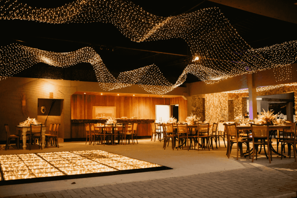 A warmly lit event space adorned with cascading string lights and floral centerpieces sets the stage for an upscale gathering. Wooden tables and chairs surround a glowing square floor feature, evoking the immersive elegance of fine dining environments—where atmosphere, design, and hospitality converge, distinct from the precision-driven ethos of Michelin-starred venues.