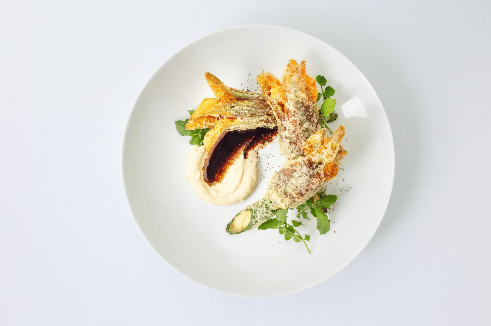 Elegant fine dining plating featuring crispy zucchini medallions fanned over a silky cauliflower purée, accented with a dark umami-rich reduction and microgreen garnish—showcasing the artistry, restraint, and flavor layering central to Singapore’s signature haute cuisine experience.