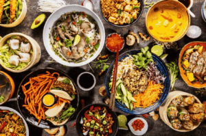 Overhead shot of colorful Singapore-style fusion dishes in small bowls on a rustic table, featuring noodles, curry, dumplings, and fresh herbs.