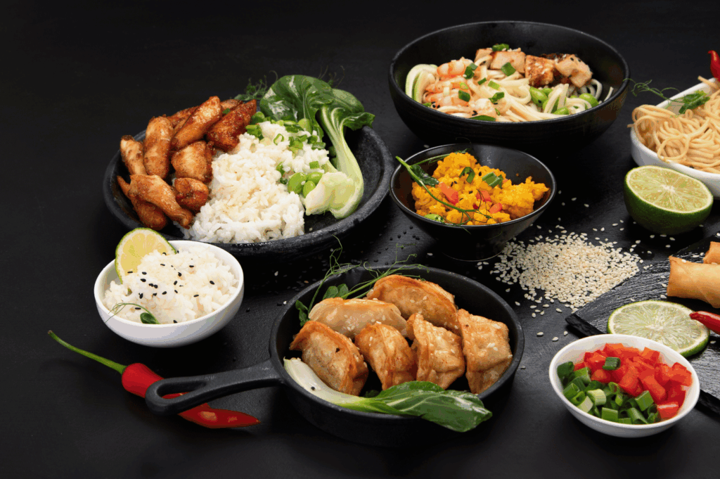 Overhead shot of vibrant Asian-inspired dishes—noodles, dumplings, skewers, and sauces—styled on a dark surface, showcasing modern fusion plating.