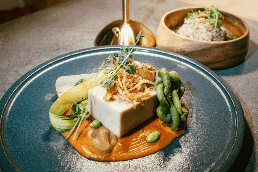 Plated vegan fine‑dining dish in Singapore: tofu cube on silky carrot purée with edamame, greens, and micro‑herbs, photographed in warm restaurant light.