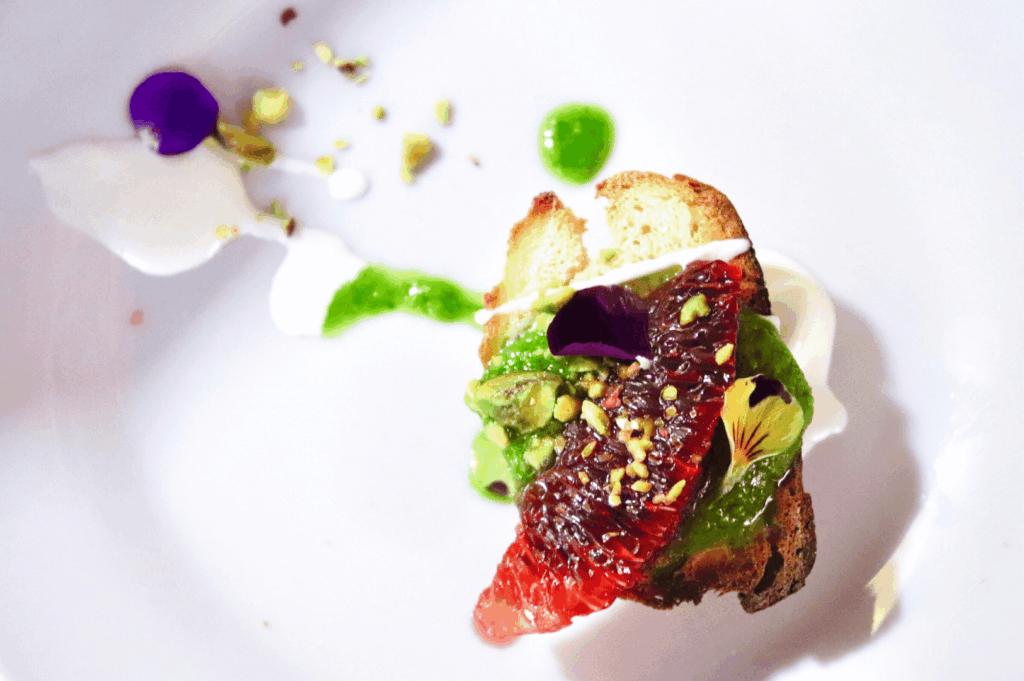 Plated vegan fine-dining starter in Singapore: toasted bread with citrus, green herb purée, pistachios, and edible flowers on a white plate, styled for a Michelin-inspired feature.
