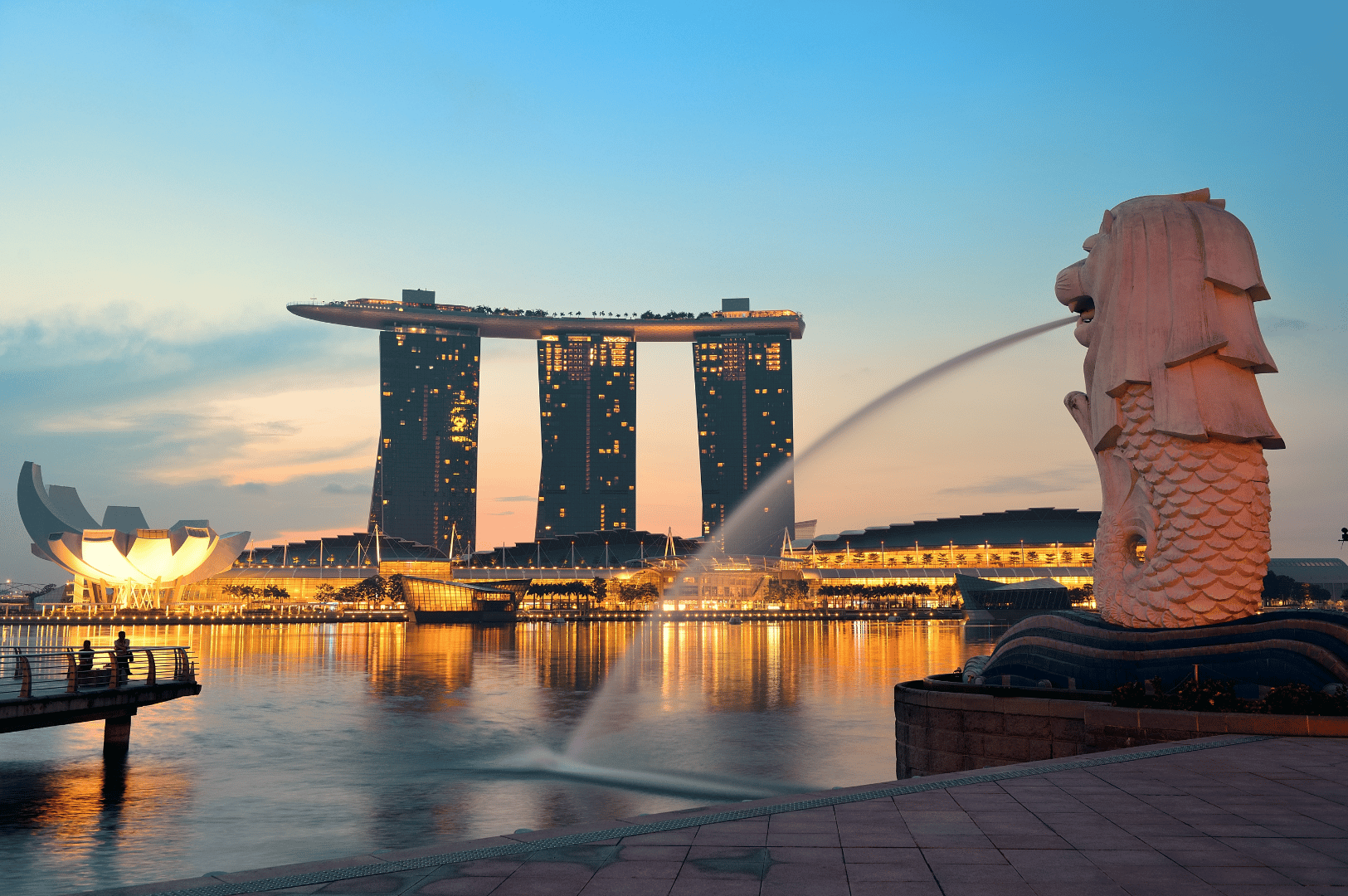 Twilight view of Marina Bay, Singapore, featuring the iconic Merlion spouting water in the foreground, with Marina Bay Sands and the lotus-shaped ArtScience Museum illuminated across the bay. Reflections ripple across the water, evoking the city's blend of architectural grandeur and waterfront serenity—an atmospheric prelude to Singapore’s most scenic dining destinations from the CBD to Sentosa.
