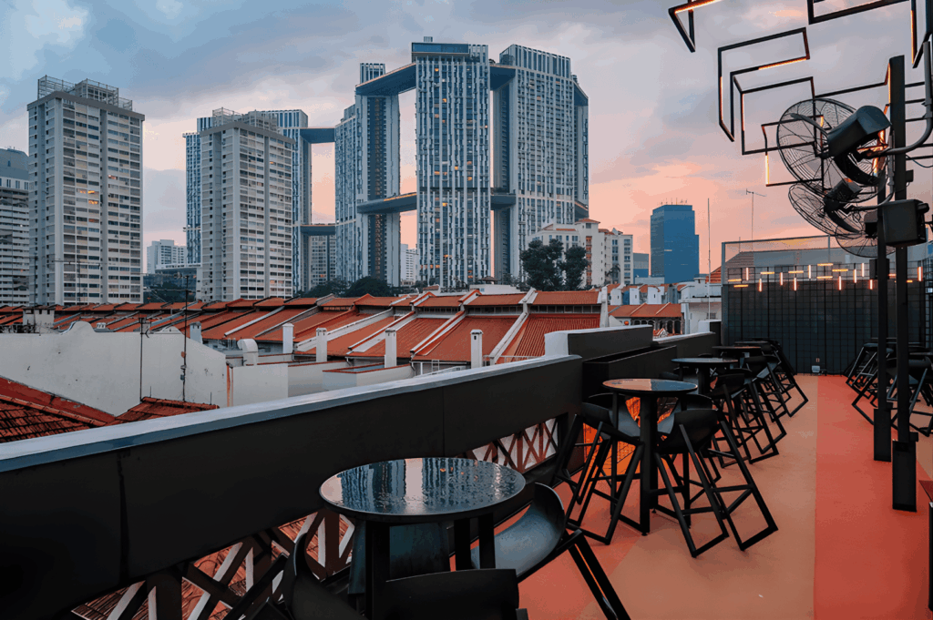 Rooftop bar overlooking Singapore’s skyline at golden hour, with sleek black tables and high chairs facing a panorama of heritage rooftops and futuristic towers. The view includes skybridged high-rises and warm-hued clouds, blending old-world charm with urban sophistication—an inviting vantage point in the city’s scenic dining journey from the CBD to Sentosa.