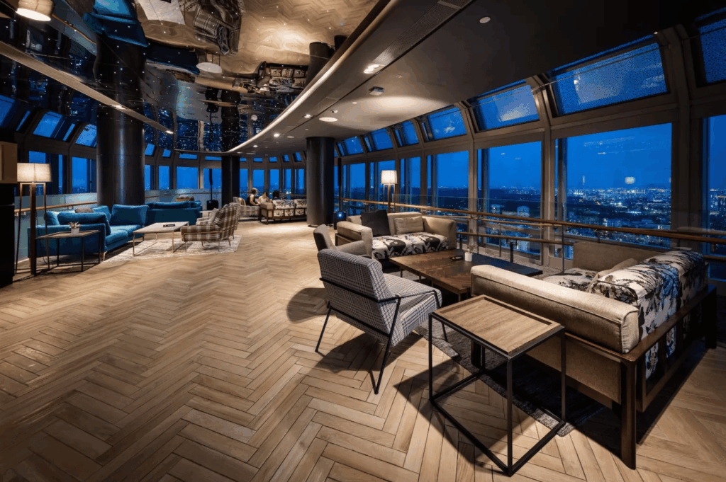 Upscale lounge in a high-rise with plush seating and herringbone wood floors, framed by floor-to-ceiling windows revealing Singapore’s city lights at dusk. Reflective ceiling panels and ambient lighting create a serene, elevated atmosphere—an intimate pause in the city’s scenic dining journey from the CBD to Sentosa.