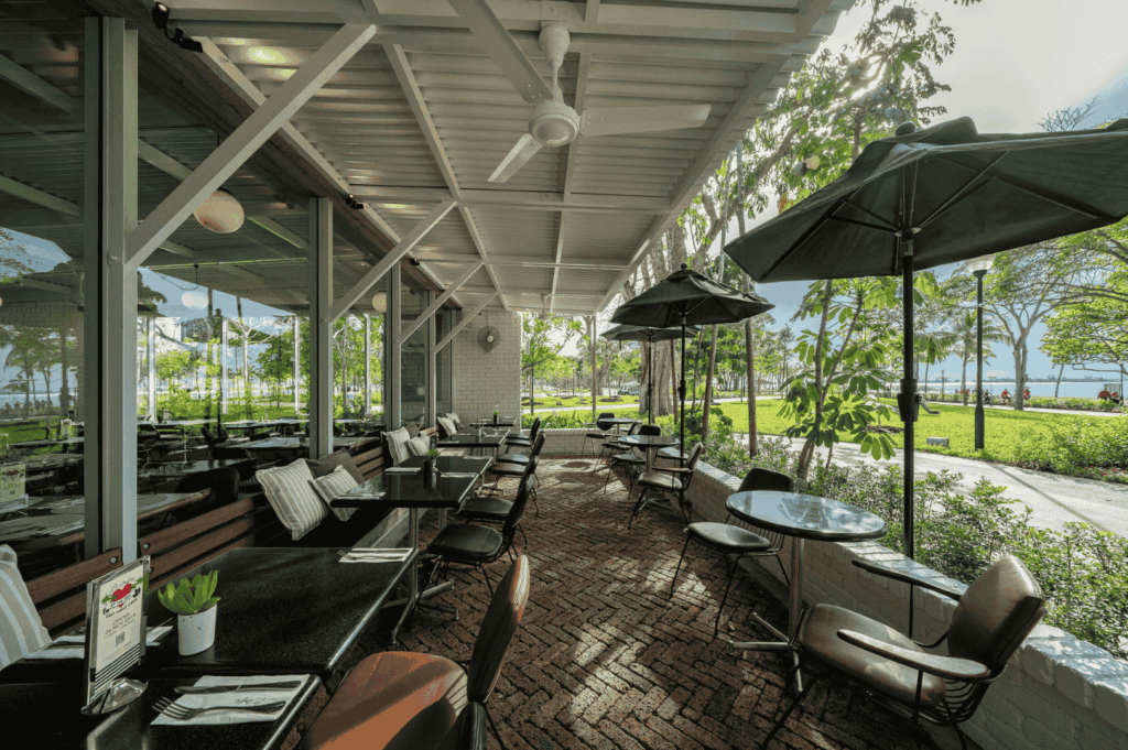 Covered patio dining area with black tables and cushioned chairs set against a red brick herringbone floor, opening onto a lush garden and distant water views. Ceiling fans and potted plants add tropical charm—an open-air retreat in Singapore’s scenic dining journey from the CBD to Sentosa.