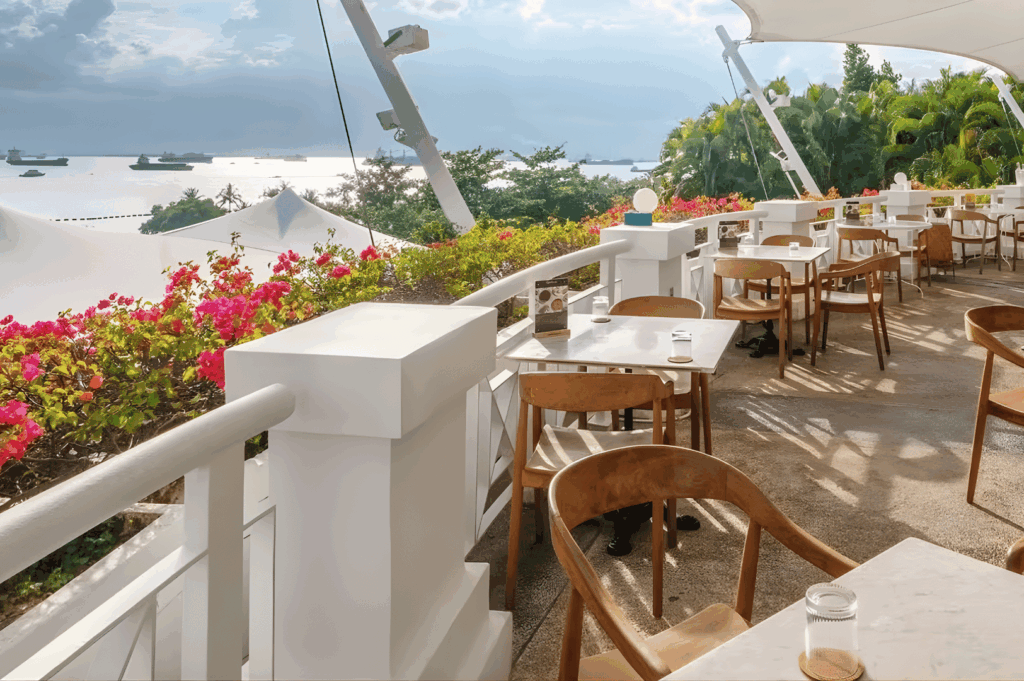 Terrace dining area with white tables and wooden chairs framed by vibrant bougainvillea and ocean views dotted with ships. White canopies and lush greenery offer shade and tranquility—an idyllic coastal finale to Singapore’s scenic dining journey from the CBD to Sentosa.