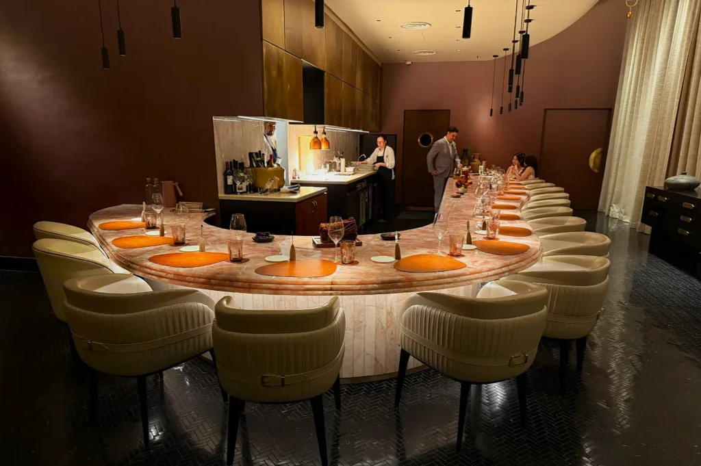 Elegant, curved marble bar with plush white chairs set for dining, warm lighting, and staff preparing drinks, creating a sophisticated ambiance.