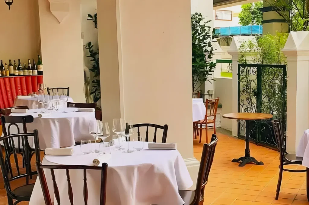 Elegant outdoor restaurant with round tables covered in white cloths, set with glassware and napkins. Warm ambiance, green plants, and decorative bottles.