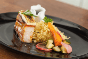 Elegant black plate showcasing a multicultural Singapore fusion dish: crispy caramelized pork belly topped with a delicate cheese sliver and parsley, paired with fragrant pilaf-style rice and a vibrant medley of pickled vegetables—radish, cucumber, carrot, and onion. A white sauce pitcher in the background hints at layered flavor traditions. The composition evokes the harmony of Chinese, Malay, Indian, and Peranakan culinary influences, celebrating Singapore’s rich cultural tapestry through modern plating and bold flavor contrasts.