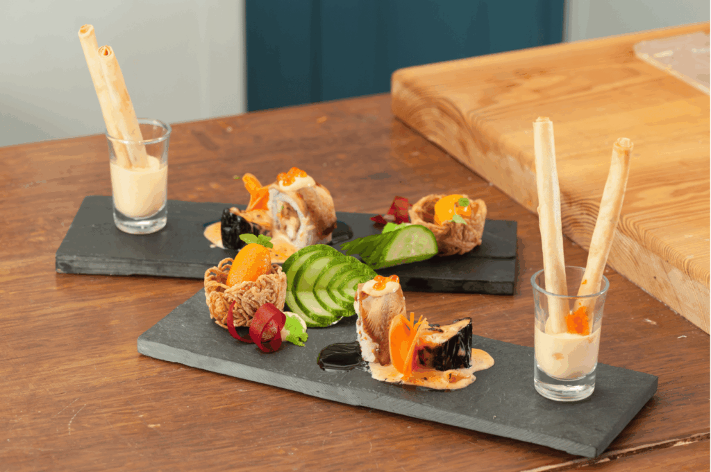 Artfully plated Singapore fusion appetizers on black slate, featuring rolled meat slices with savory sauce, fan-shaped cucumber ribbons, and a crisp vegetable basket alongside a mysterious black gelatin element. Garnishes of vibrant carrot and red pepper add visual punch, while creamy dipping sauces in clear glasses with vertical breadsticks evoke Western fine dining flair. The composition reflects Singapore’s multicultural palate—melding Chinese, Malay, Indian, and European influences into a visually striking, texturally diverse culinary tableau.