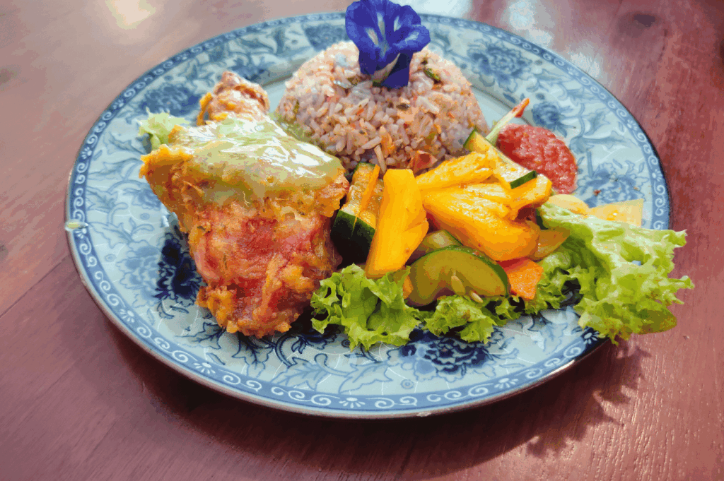 Decorative blue-and-white plate featuring a vibrant Singapore fusion entrée: golden-crusted chicken topped with herbaceous green sauce, paired with dome-shaped vegetable rice crowned by a purple edible flower. A tropical salad of mango, cucumber, lettuce, and tomato adds freshness and color. Set on a wooden surface, the dish blends Southeast Asian ingredients with Western plating finesse, reflecting Singapore’s multicultural spirit through balanced textures, bold hues, and thoughtful presentation.