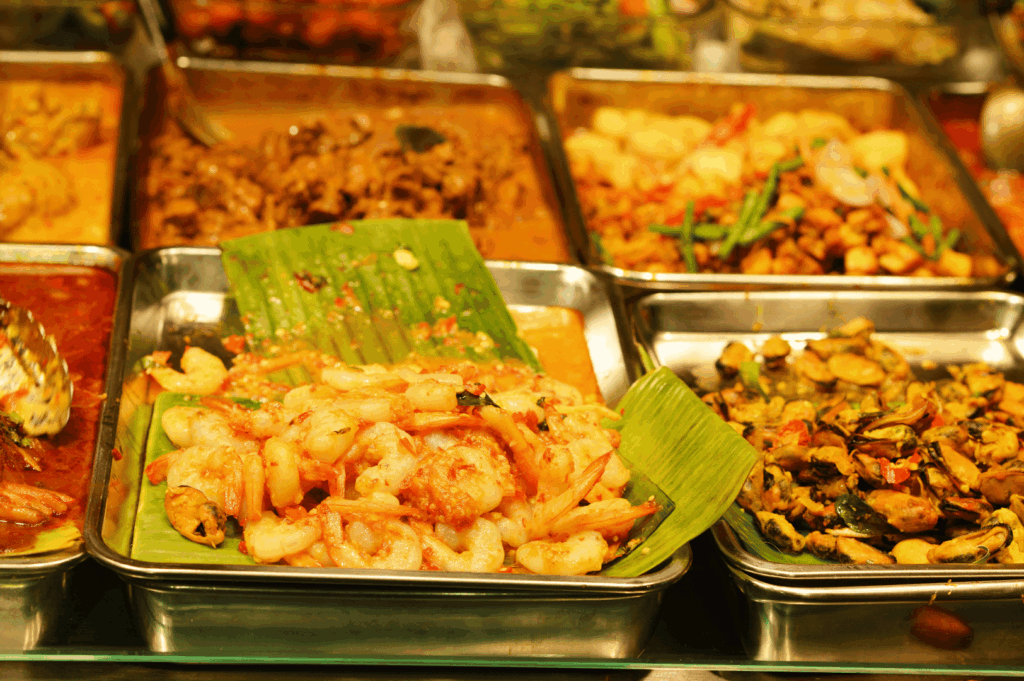 Vibrant array of cooked dishes displayed in metal trays at a buffet-style setting, featuring banana leaf–lined shrimp, stir-fried vegetables, and richly sauced curries. The colorful spread includes meats, seafood, and plant-based options, reflecting the communal dining traditions of Singapore’s hawker culture. The visual abundance and diversity of flavors pay homage to the nation’s multicultural roots—Chinese, Malay, Indian, and Eurasian—where everyday meals become a celebration of shared heritage.