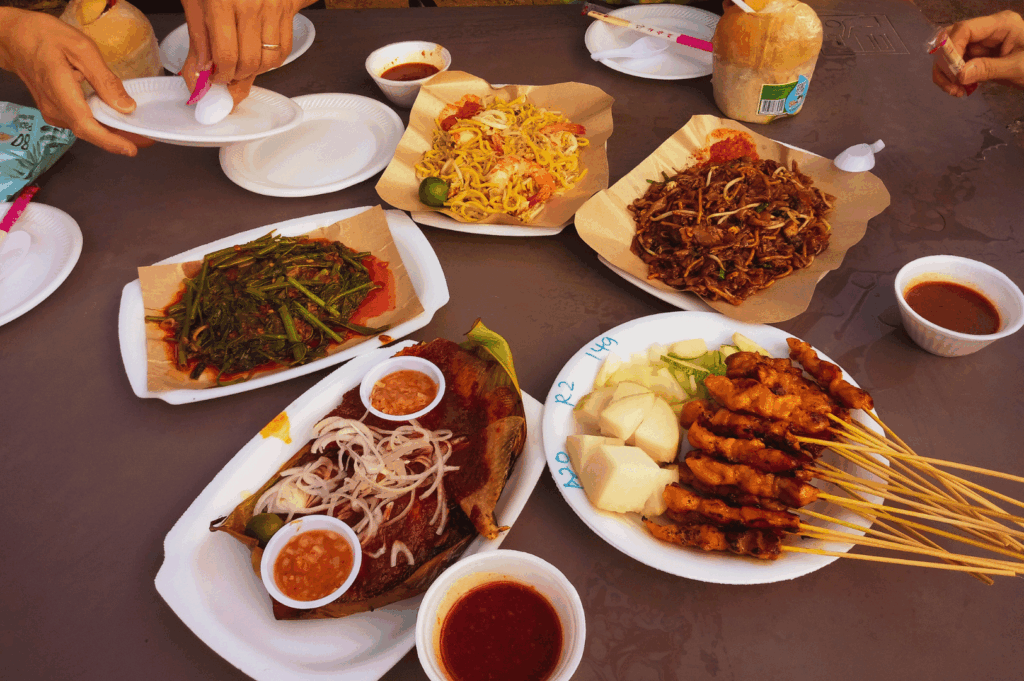 Casual street-style spread of Singapore fusion dishes arranged on a shared table: grilled chicken satay with cucumber and dipping sauce, grilled fish with onions, stir-fried morning glory, and assorted noodle bowls with vegetables and meat. Coconut drink and plastic utensils add everyday charm, while reaching hands evoke the communal joy of dining. The vibrant mix of Malay, Chinese, and Indian flavors reflects Singapore’s multicultural soul, where food is both heritage and connection.