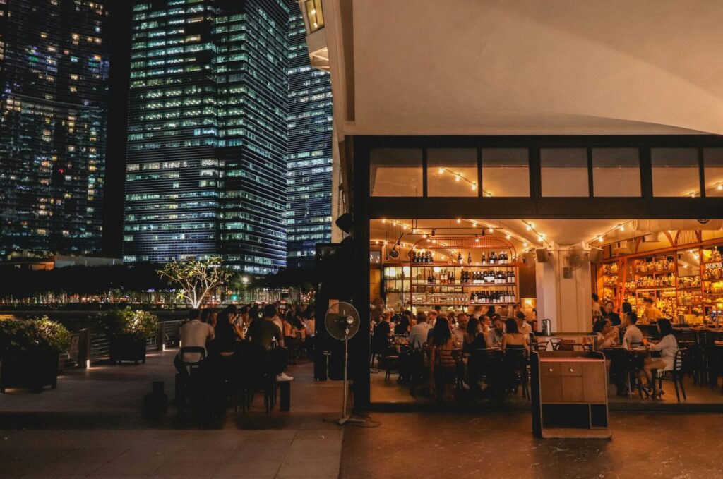A bustling restaurant scene with diners at tables, set against the backdrop of tall buildings in Singapore's luxury district.