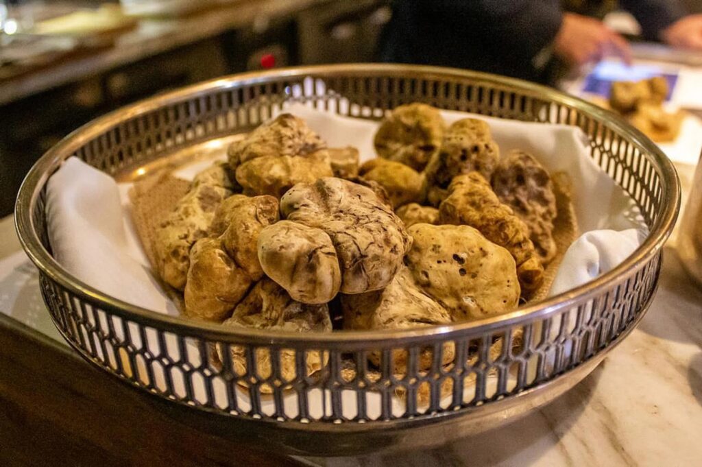 A basket filled with large, textured white truffles on a white napkin, placed on a marble countertop. Soft, ambient lighting suggests a cozy setting.