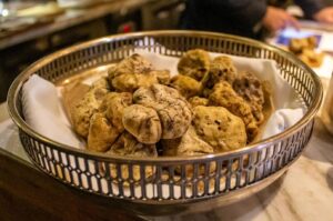 A basket filled with large, textured white truffles on a white napkin, placed on a marble countertop. Soft, ambient lighting suggests a cozy setting.