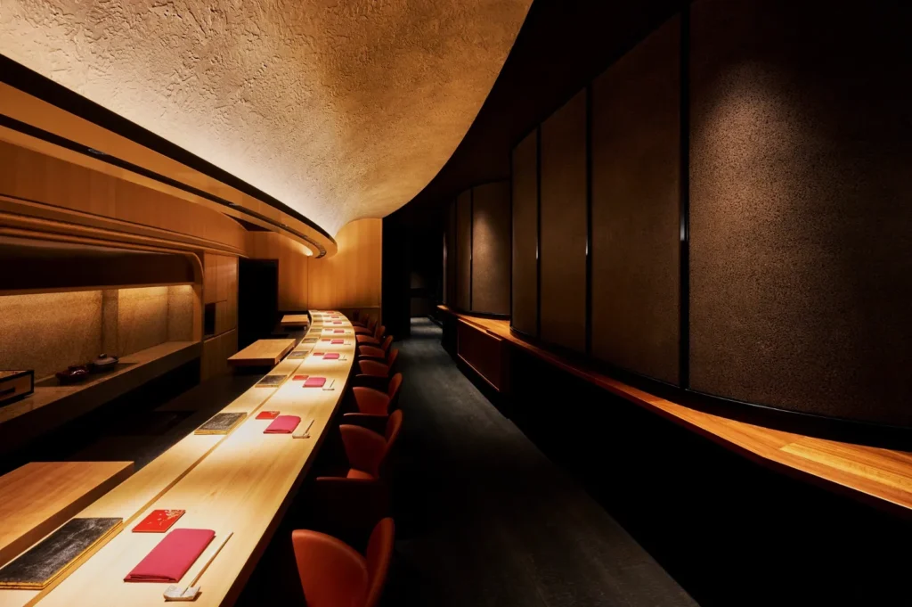 Elegant, dimly lit sushi bar with a long wooden counter, red chairs, and curved ceiling. Warm, inviting ambiance with subtle textures and muted colors.