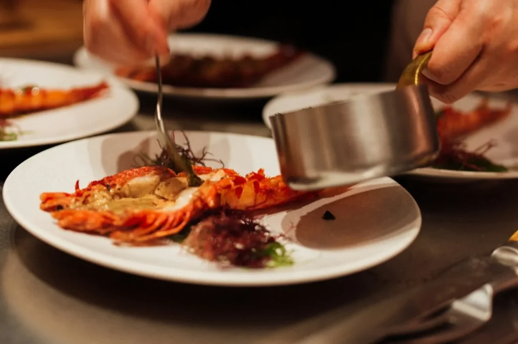 Chef's hands artfully plating a dish with lobster and seaweed, adding sauce from a small pot. The scene conveys a sense of gourmet cuisine.