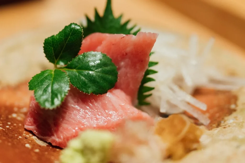 Close-up of a sushi plate featuring fresh pink tuna sashimi garnished with green leaves. Shredded daikon and wasabi add texture and color contrast.