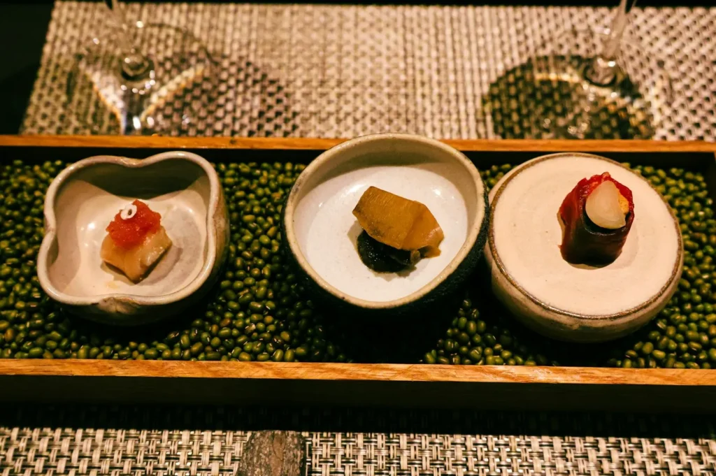 Three artistic dishes presented in small bowls rest on a bed of green beans. The setting exudes elegance with a textured placemat and wine glasses.