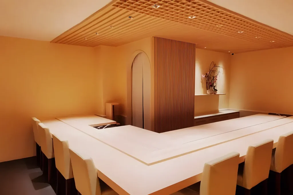 Minimalist dining area with a U-shaped wooden counter surrounded by cream chairs. Soft lighting, a slatted ceiling, and a flower arrangement create a serene ambiance.