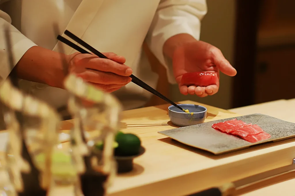 A chef in a white coat prepares sushi, holding a piece of raw tuna with chopsticks over a blue dish. Slices are on a gray plate, exuding precision and calm.