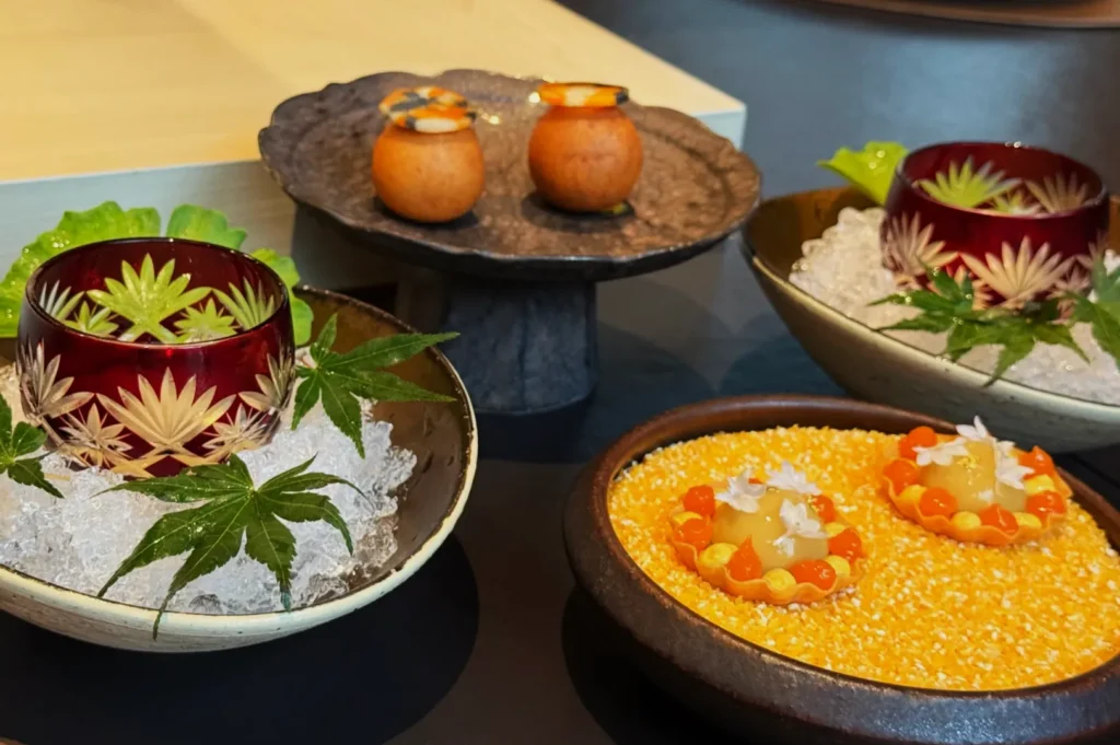 A gourmet dish with intricate plating features red bowls on ice with maple leaves, stone bowls with creamy desserts, and vibrant yellow accents. Elegant and artistic.