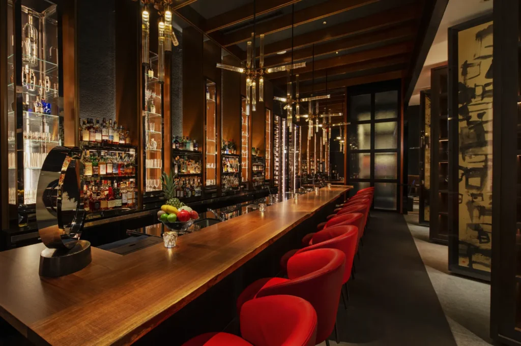 Elegant bar interior with a long wooden countertop, lined with plush red chairs. Warm lighting from modern pendant lamps creates a cozy, sophisticated ambiance.
