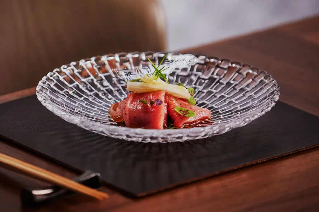 Delicate slices of tuna sashimi garnished with herbs and daikon, served on a textured glass plate. A serene dining setting with wooden chopsticks.