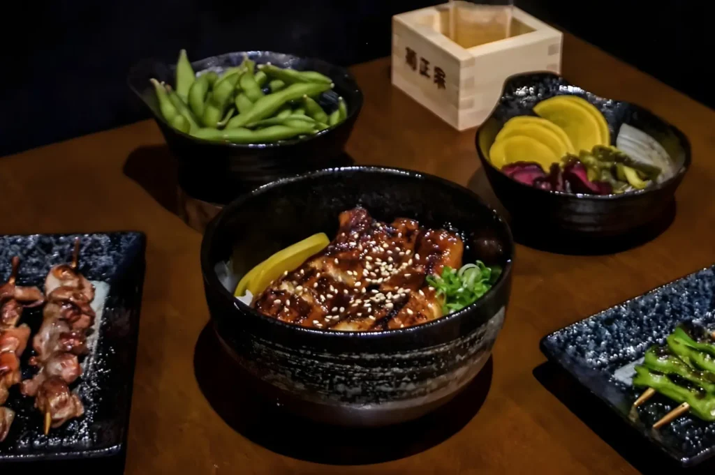 A table with Japanese cuisine, featuring a glazed chicken bowl with sesame seeds, pickled vegetables, edamame, a wooden sake box, and skewers, creating a cozy dining atmosphere.