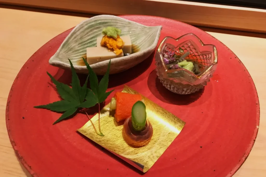 A delicate Japanese appetizer arrangement on a red plate includes assorted items in small dishes, garnished with leaves. The presentation is artistic and refined.