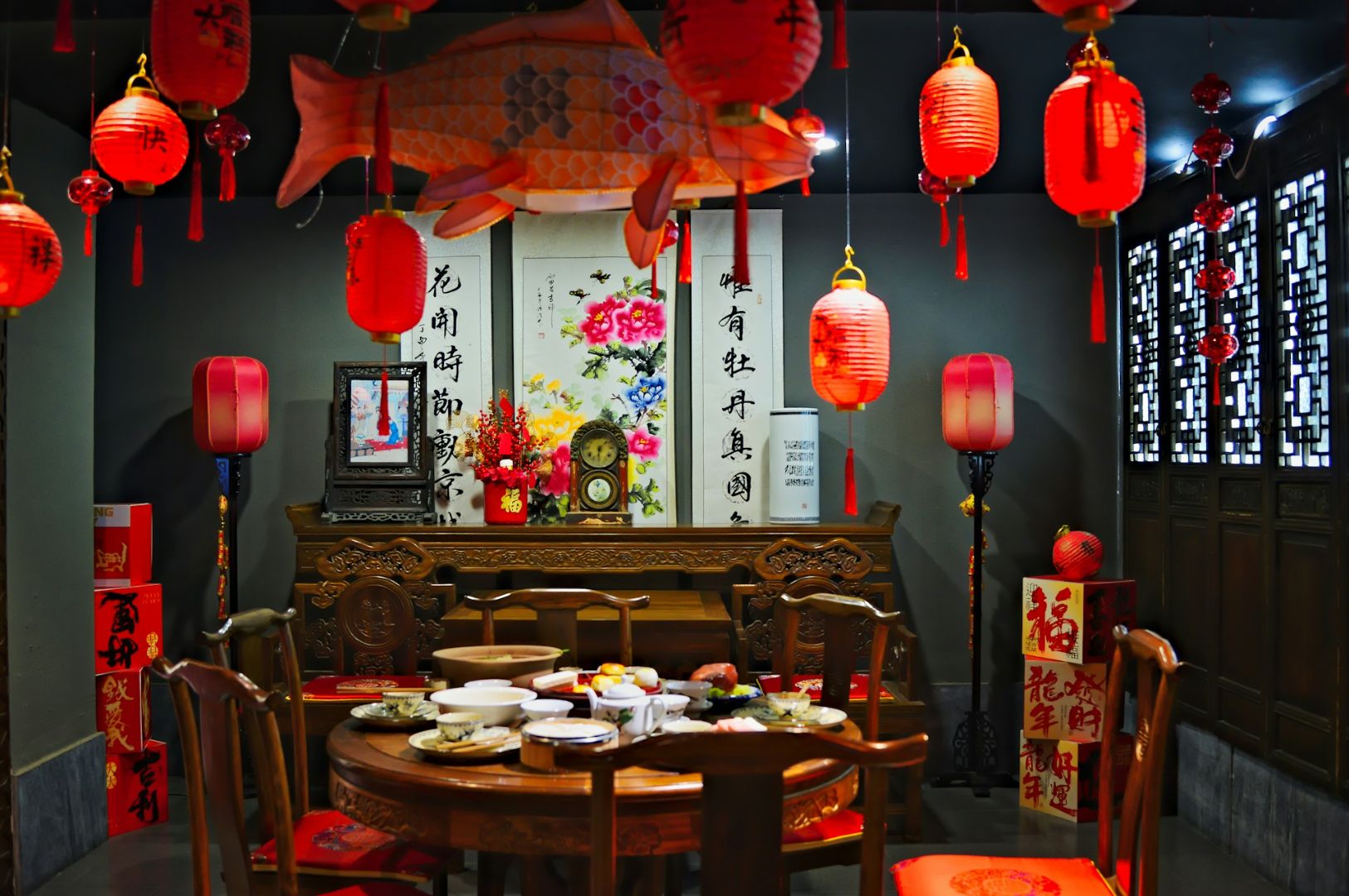 A traditional Chinese dining room decorated for a celebration. Red lanterns and a fish ornament hang above a table filled with dishes, surrounded by ornate chairs. Chinese calligraphy and floral artwork adorn the walls, creating a festive and warm atmosphere.