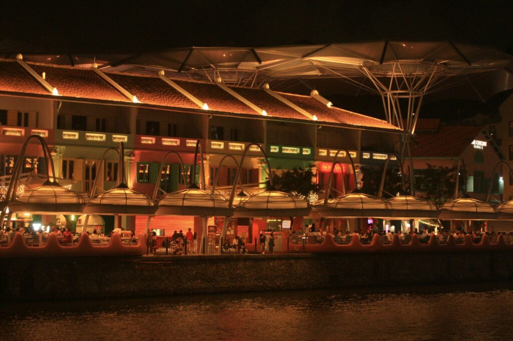 Illuminated riverside building at night with a bustling atmosphere. Cafés and restaurants are visible under warm lights, with a reflective water surface.