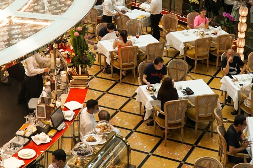 Elegant restaurant scene from above with diners at white-clothed tables, waiters serving, and a pastry-laden counter. The atmosphere is lively and sophisticated.
