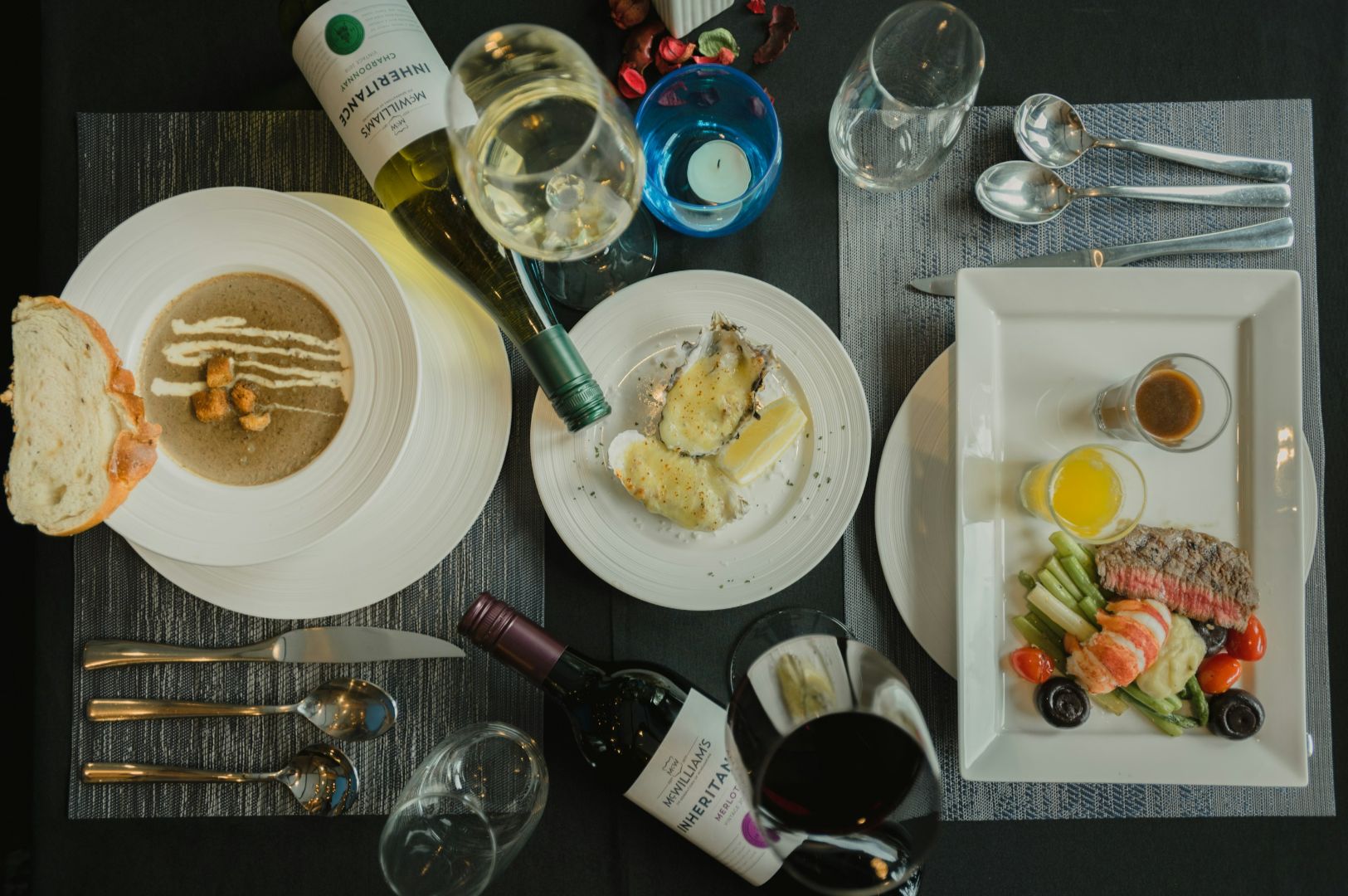 A sophisticated dining table setting with creamy soup, oysters, and a dish of steak and vegetables. Wine bottles and glasses add an elegant touch.