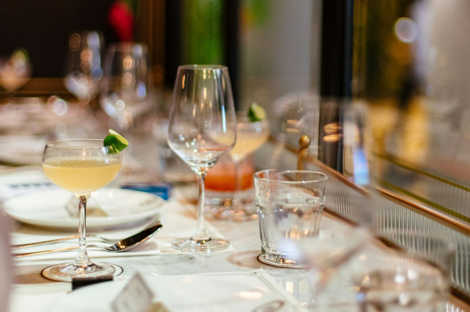 Elegant dining table set with cocktails, water glasses, and empty wine glasses on a white tablecloth. Soft, warm lighting creates a cozy ambiance.