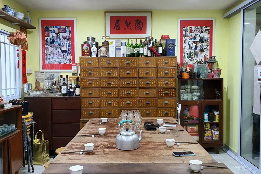 A rustic Chinese private dining room featuring a traditional wooden herbal medicine cabinet, a long wooden table set with tea cups, and walls decorated with Chinese calligraphy and photo collages.