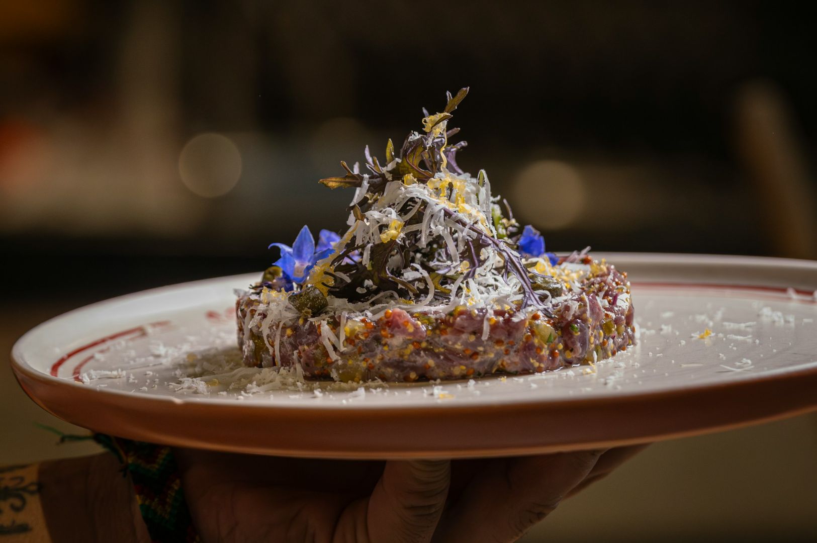 A plate features elegantly arranged tartare topped with grated cheese, edible flowers, and delicate greens, creating a gourmet, artistic presentation.