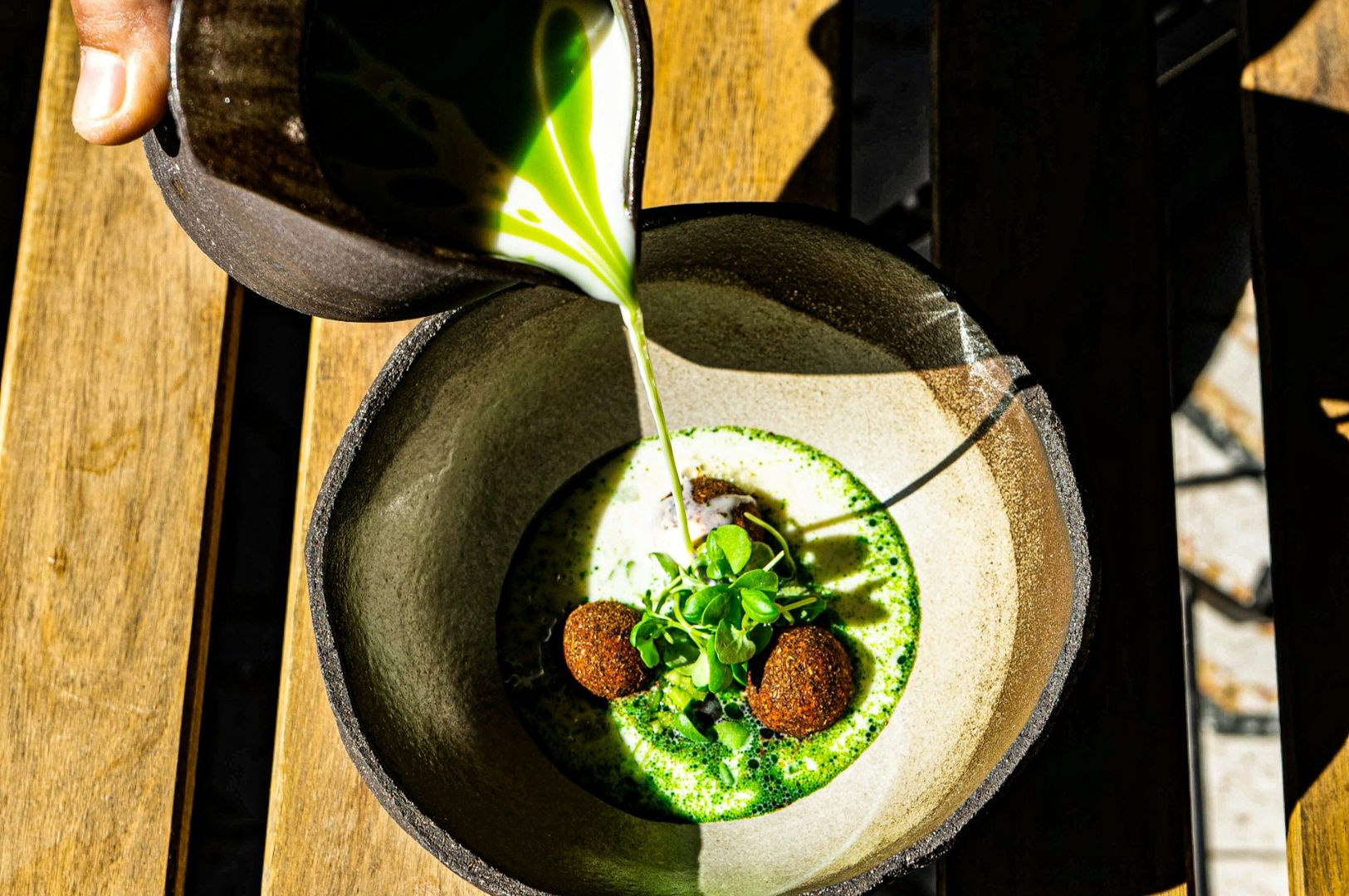 A hand pours green soup into a rustic bowl on a wooden table, surrounding two brown croquettes and fresh greens. The scene feels fresh and inviting.