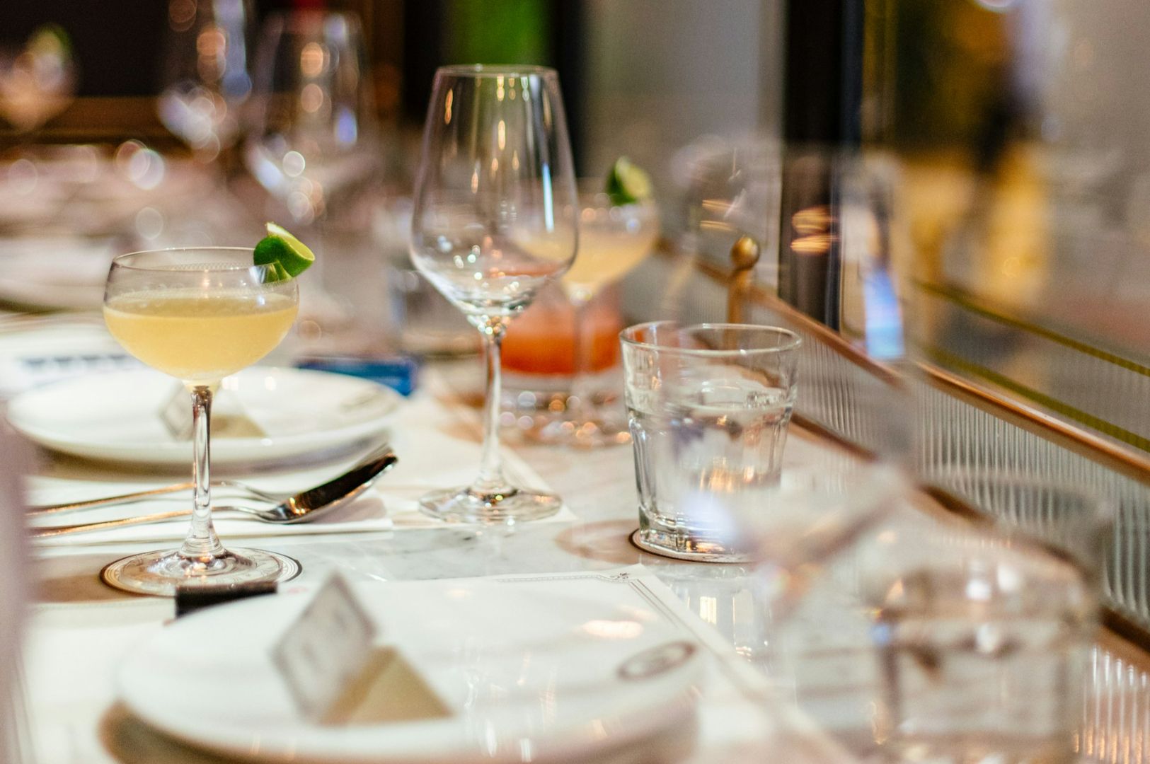 Elegant table setting with cocktail glasses garnished with lime, wine glasses, and water tumblers on a white tablecloth. Soft, inviting ambiance.