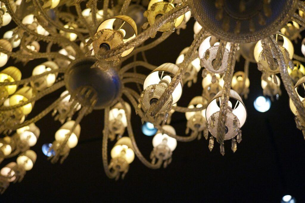 A close-up of an ornate chandelier with illuminated bulbs casting a warm, elegant glow. Curved, intricate arms create a luxurious, vintage ambiance
