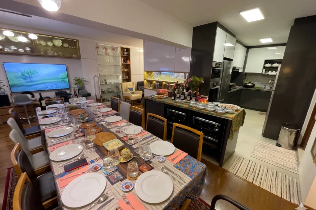 An elegant open-concept home dining area with a long table set for a multi-course meal, featuring batik table runners and a view of a modern kitchen and wine coolers.
