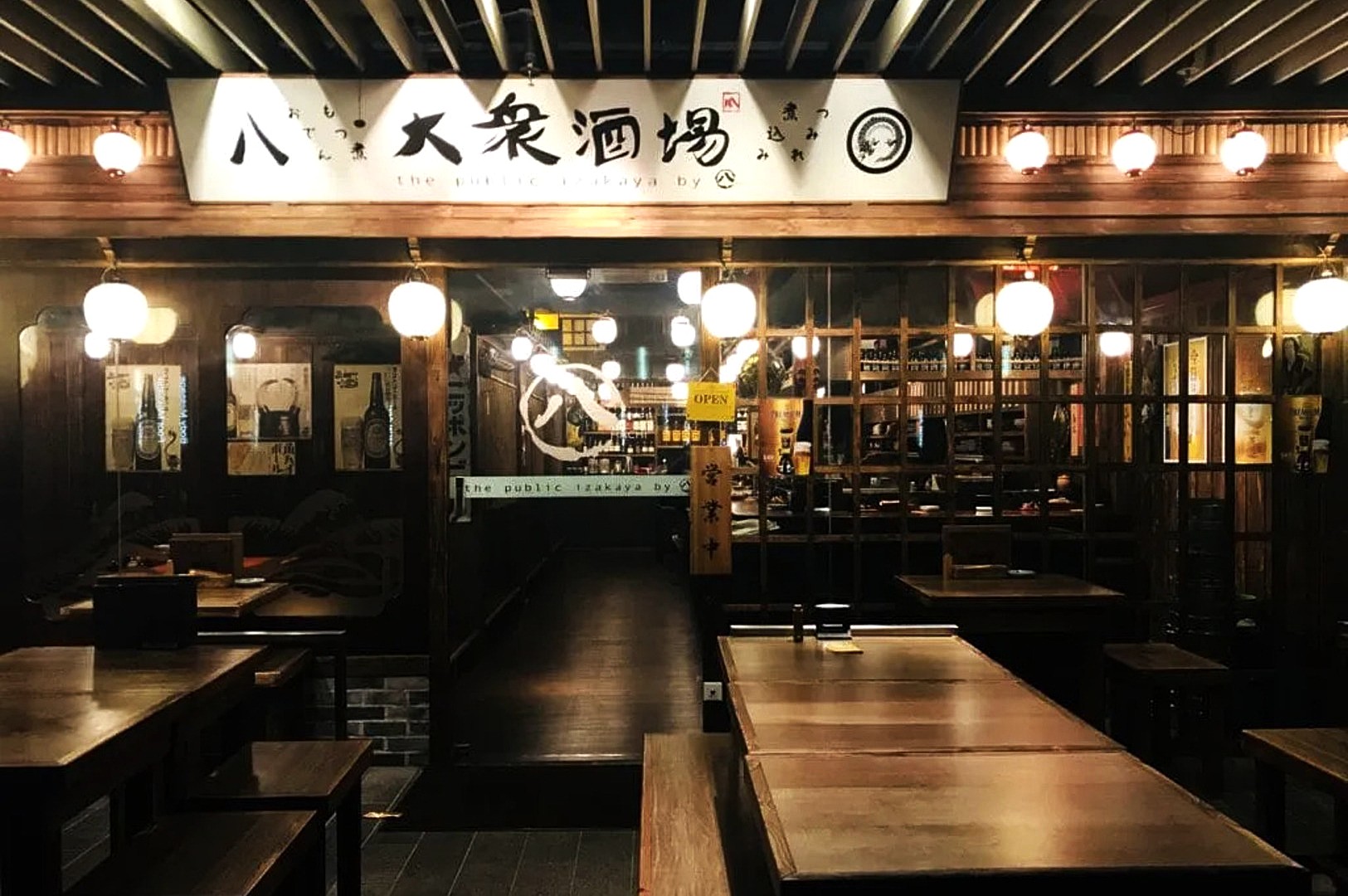 Entrance of a cozy Japanese izakaya, warmly lit with hanging lanterns. Wooden tables and interior evoke a traditional, inviting atmosphere.