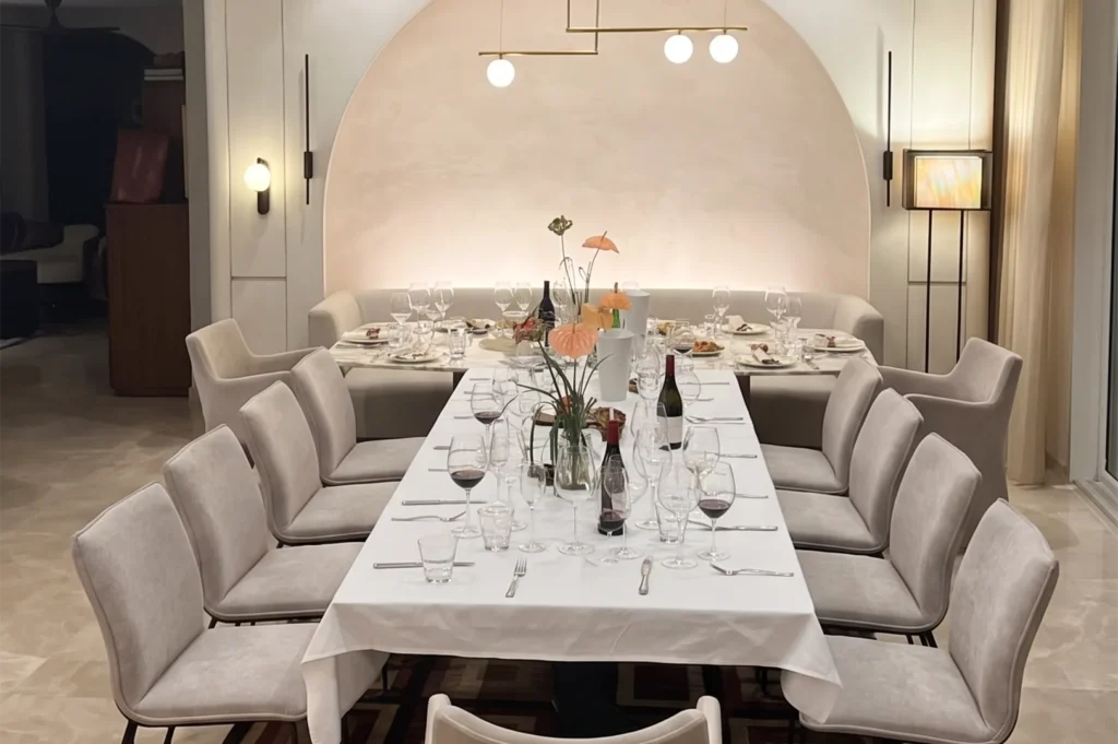A high-end minimalist dining room with a long white-clothed table, velvet chairs, and a large recessed arched wall feature with soft ambient lighting.