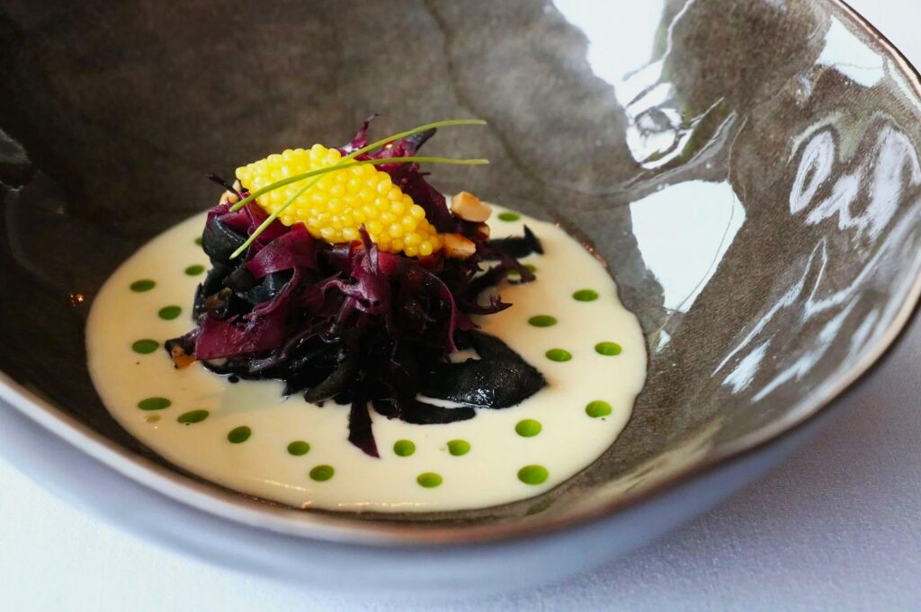 Gourmet dish with vibrant yellow pearls, purple shreds, and greens over creamy sauce with green dots. Presented in a reflective bowl, evoking elegance.
