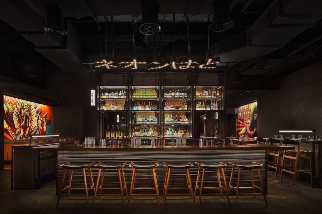 Sophisticated bar interior with dim lighting, a central shelf displaying colorful bottles, wooden stools, and vibrant murals on the dark walls. Cozy and inviting.